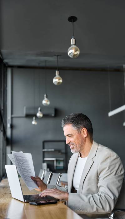 Mid aged happy professional business man company executive manager wearing suit sitting at desk in office working checking bills corporate financial accounting document using laptop computer. Vertical